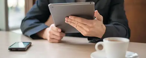 Man sitting at a booth looking at a tablet with a phone and coffee cup on the table.